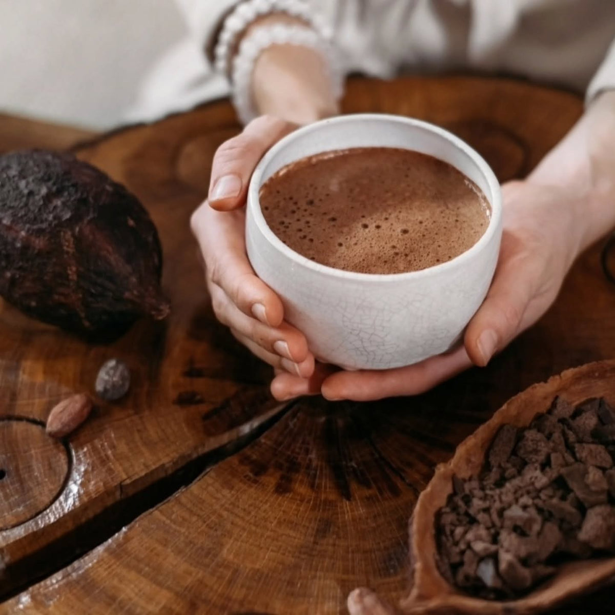 Hands holding a cup of cacao over a wooden table with cacao beans and chopped cacao