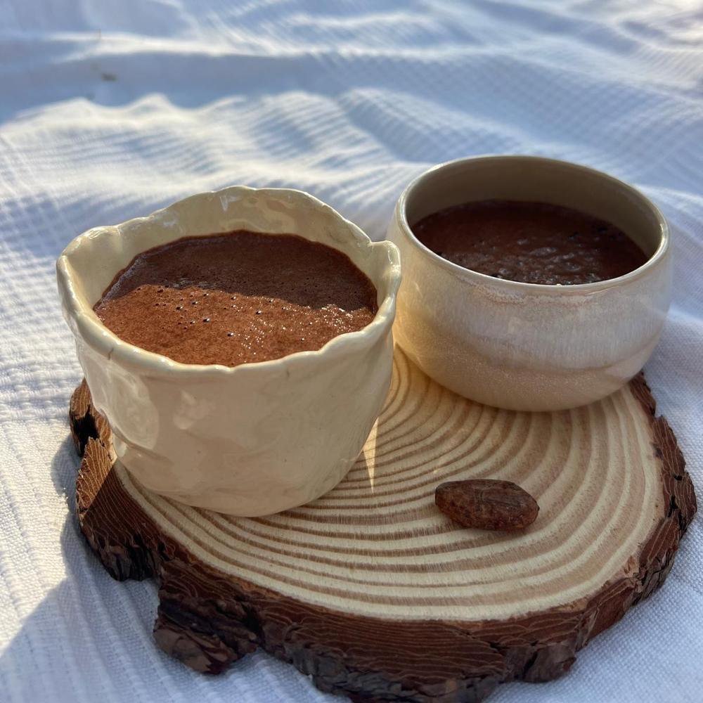 Two ceramic bowls with ceremonial grade cacao drinks on a wooden coaster on a white fabric background